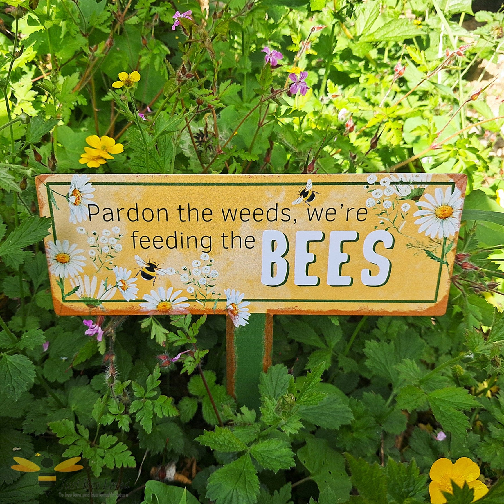 Decorative sign in a garden with flowers and plants, featuring text 'Pardon the weeds, we're feeding the BEES'.