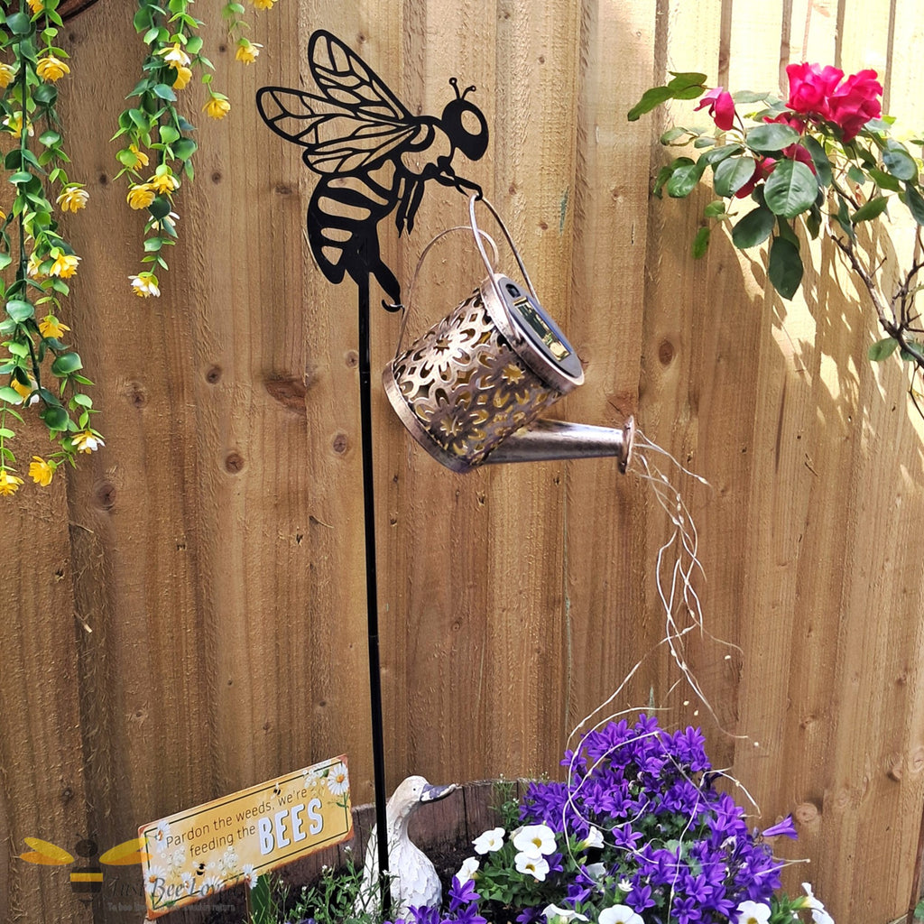 Metal garden stake shaped like a bee with a solar light watering can feature and cascading string lights.