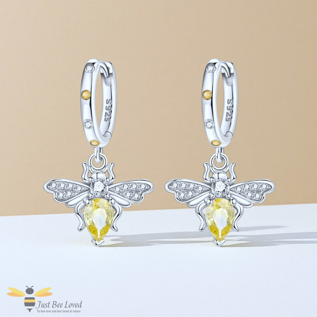 A pair of silver hoop earrings with dangling bee charms. Each bee charm features a yellow pear-cut gemstone, small clear crystals on its wings.