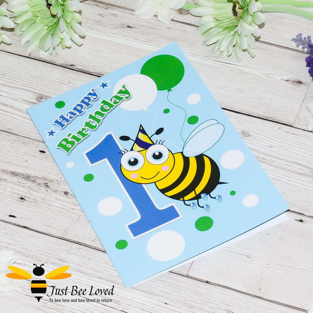 a blue birthday bee greeting card featuring a cartoon illustration of an infant bee wearing a party hat, holding white and green balloons next to the number 1.