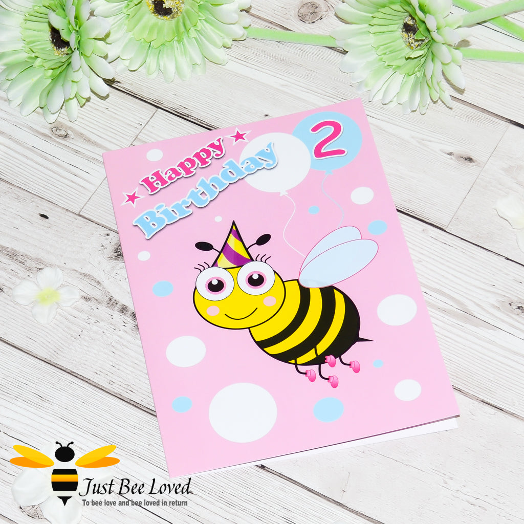 a pink birthday greeting card featuring a cartoon bee wearing a party hat, holding colourful balloons with the number 2, and 'Happy Birthday' text.