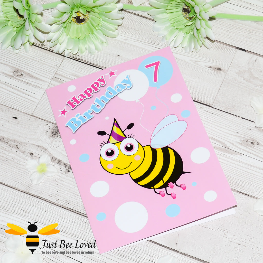 a pink birthday card with blue and white polka dots, with a cartoon illustration of a bee wearing a party hat, balloons and the number 7.