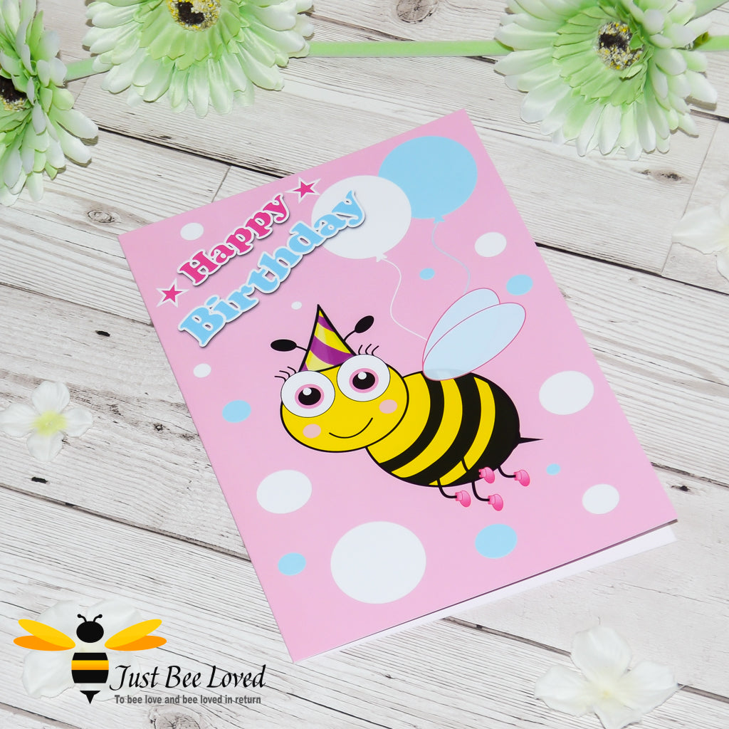 a pink birthday bee card with a cartoon illustration of a toddler bee wearing a pink hat and holding colourful balloons.