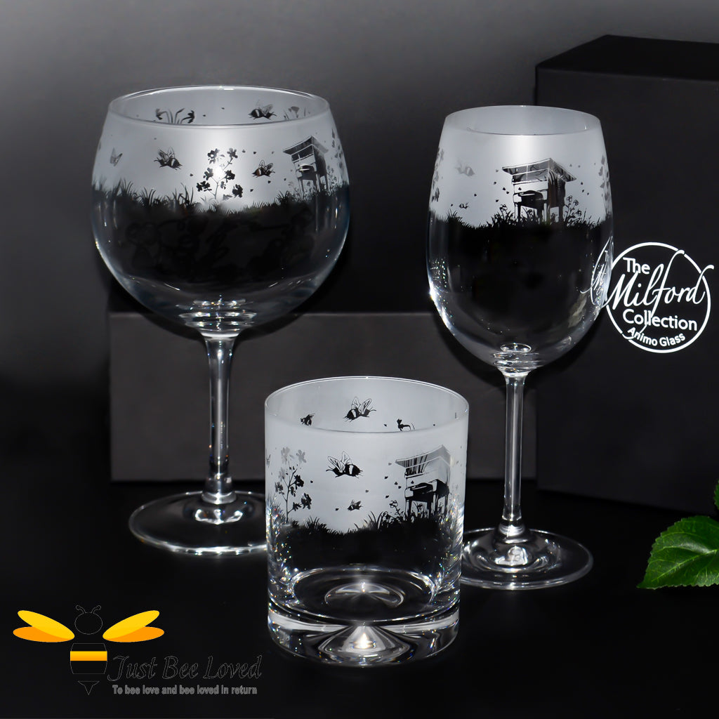 A set of three etched glass drinkware items with a frosted bee and flower design, including a whiskey tumbler, a balloon gin glass, and a stemmed wine glass.