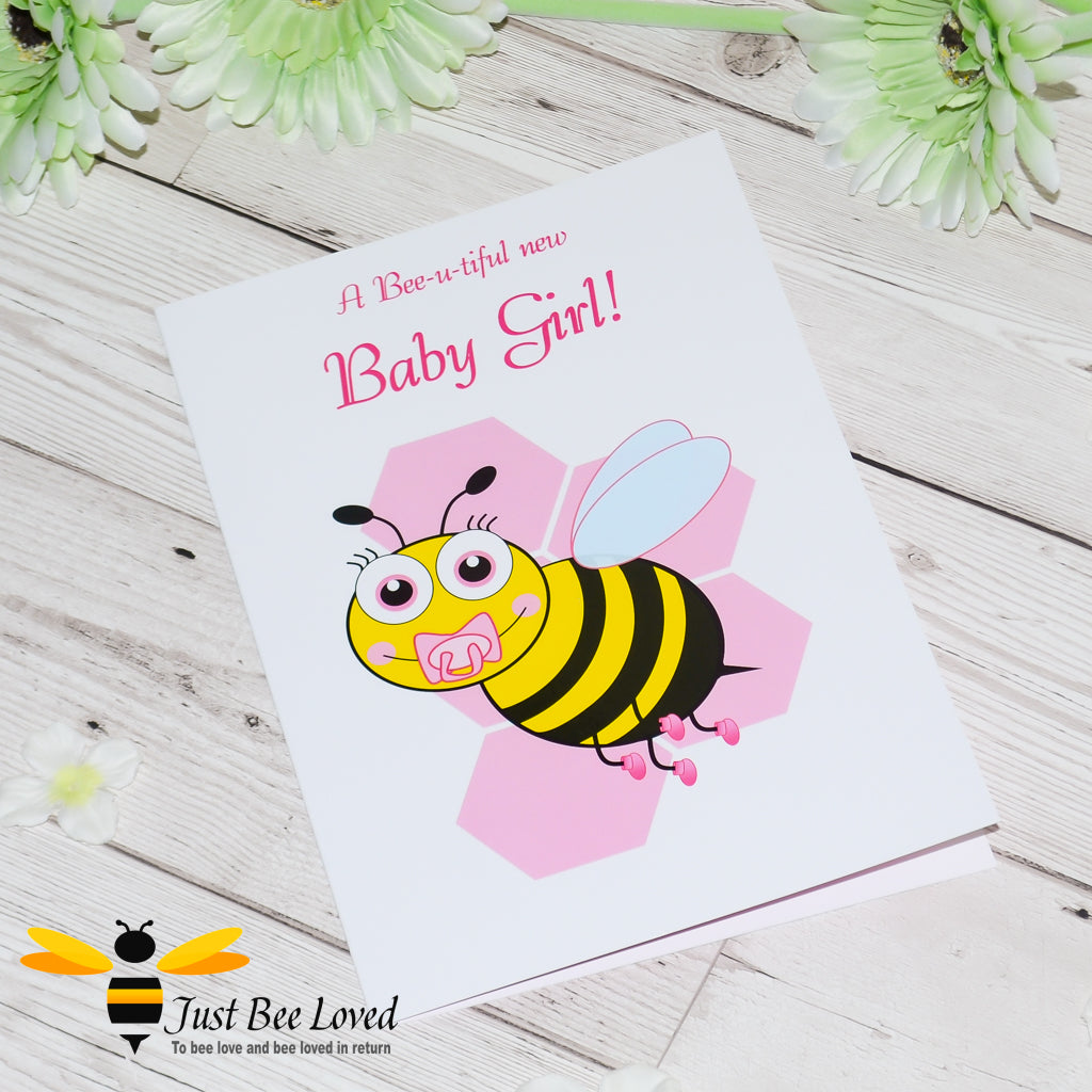 image of a new baby girl newborn bee greeting card featuring a cartoon illustration of a baby bee with a pink dummy and booties, on a honeycomb background.