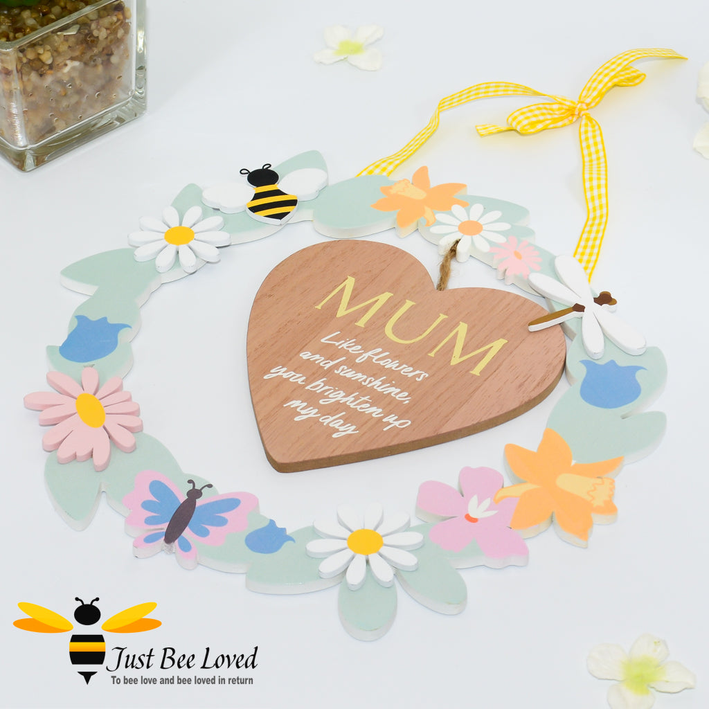 A wooden hanging plaque with a heart-shaped cut-out in the center, surrounded by colourful artificial flowers, bee motifs and the word 'MUM' inscribed on the heart.