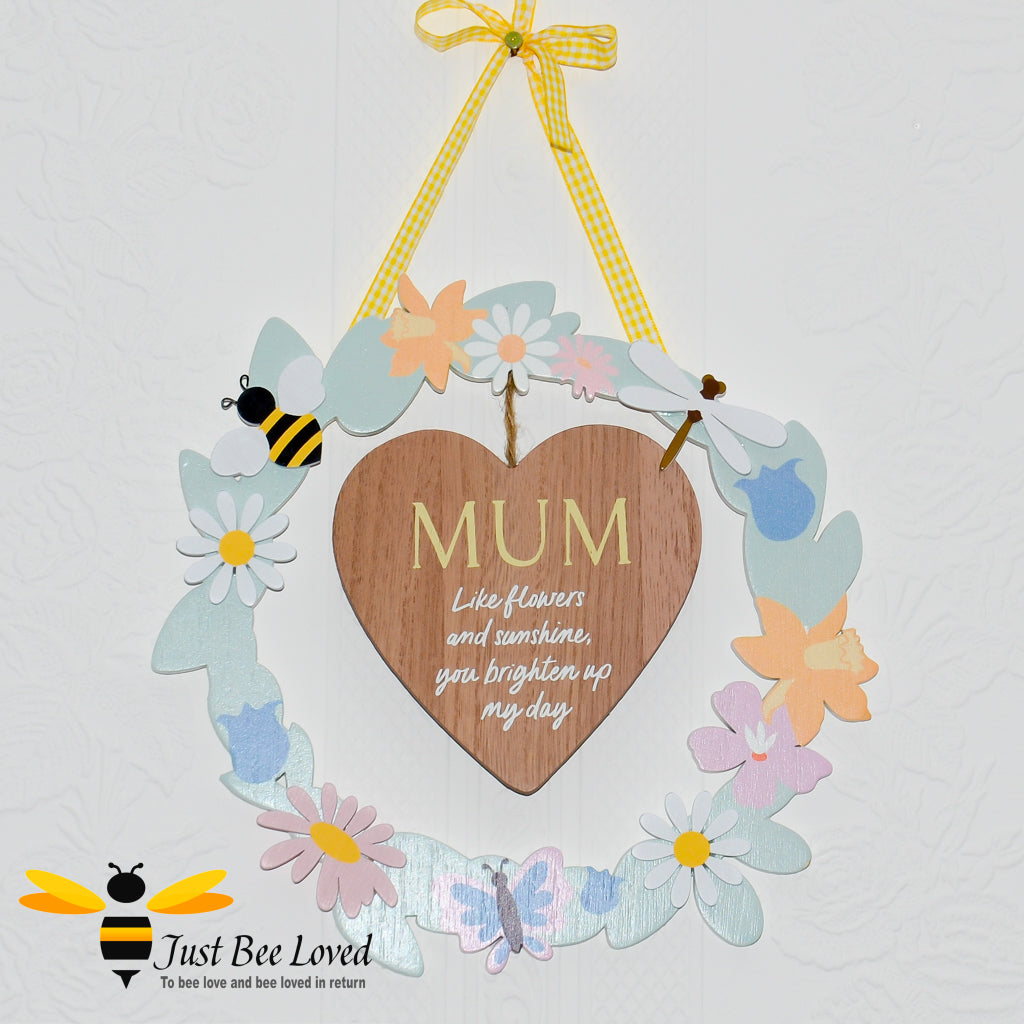 A wooden hanging plaque with a heart-shaped cut-out in the center, surrounded by colourful artificial flowers, bee motifs and the word 'MUM' inscribed on the heart.
