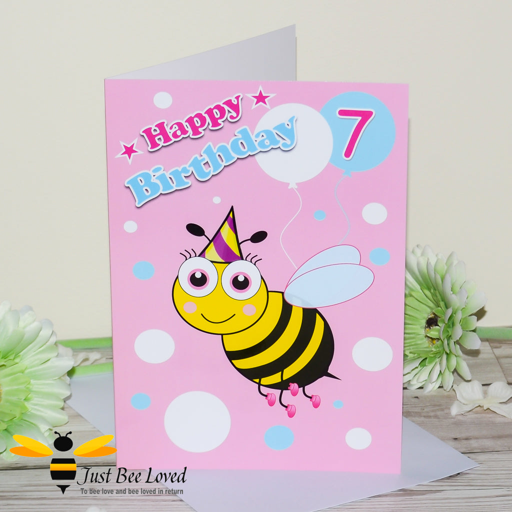 a pink birthday greeting card for girl featuring a cartoon bee wearing a party hat, balloons and the number 7.
